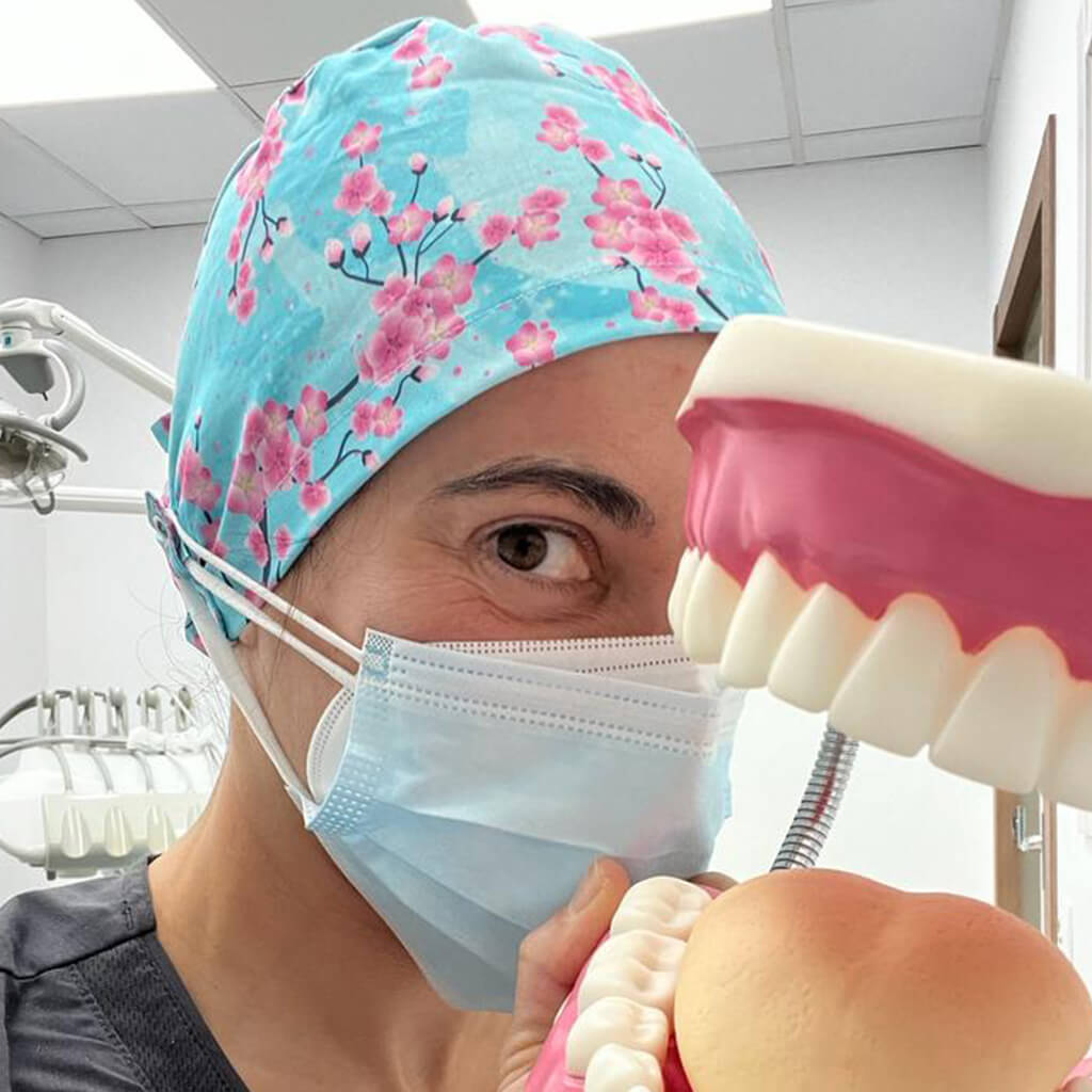 Dentista en clínica dental usando gorro quirúrgico SAKURA Robin Hat celeste con flores de cerezo rosas, mascarilla azul, modelo anatómico dental