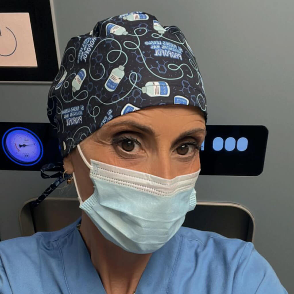 Anestesióloga en quirófano usando gorro quirúrgico ANESTESIA de Robin Hat con diseño de jeringas y propofol sobre fondo negro, llevando mascarilla durante procedimiento médico en entorno hospitalario especializado en anestesiología