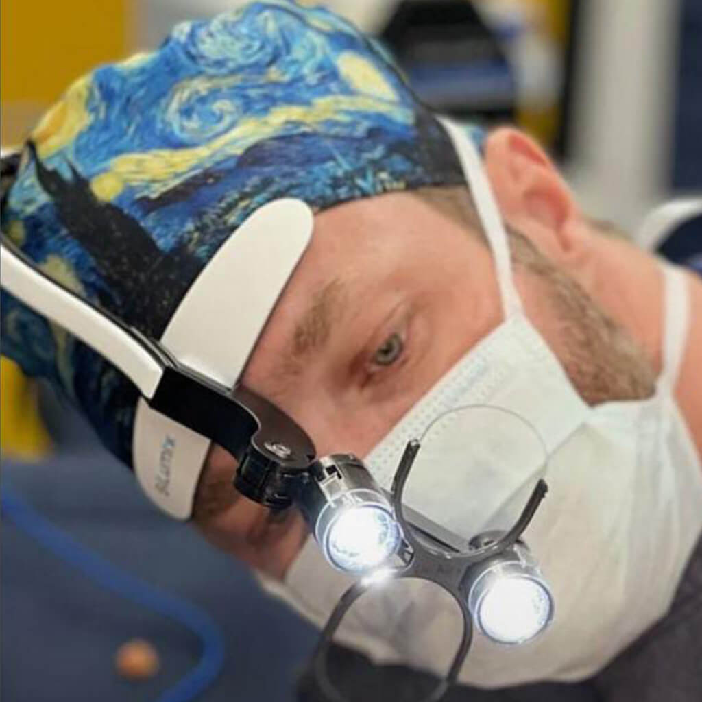 Cirujano usando gorro quirúrgico NOCHE ESTRELLADA de Robin Hat inspirado en la obra de Van Gogh con cielo en remolino y colores vibrantes, trabajando con microscopio quirúrgico y lupas durante intervención de precisión confeccionado en poliéster transpirable