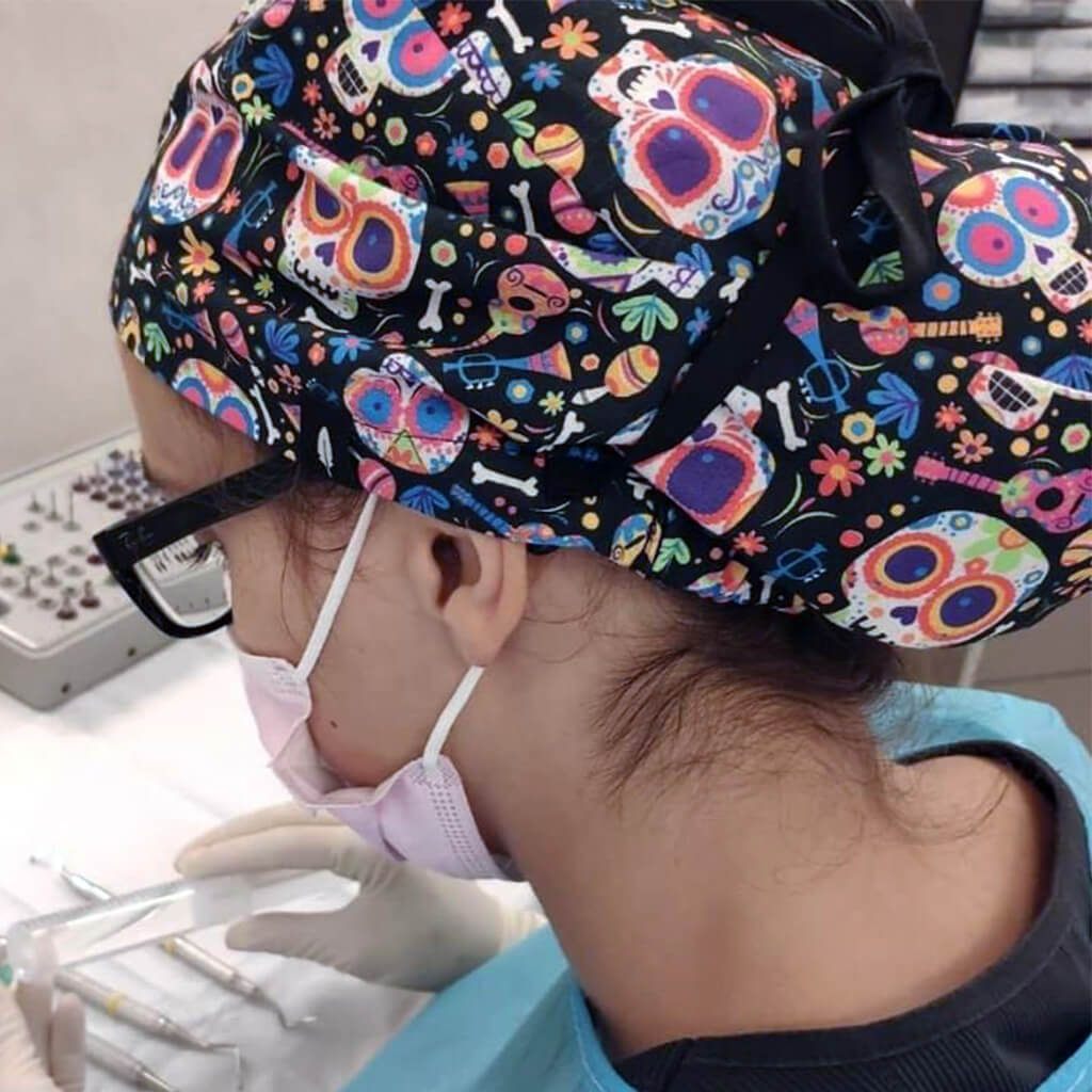 Profesional sanitario usando microscopio con gorro quirurgico Robin Hat modelo Catrina, estampado de calaveras mexicanas coloridas y flores sobre fondo negro, ideal para laboratorios y clinicas