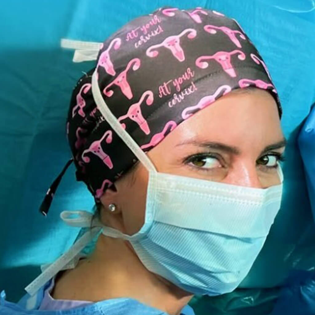 Ginecologa en sala de operaciones con gorro quirurgico Cervix de Robin Hat, estampado anatomico de uteros en tonos rosas sobre fondo negro, diseño con humor medico para especialistas en ginecologia