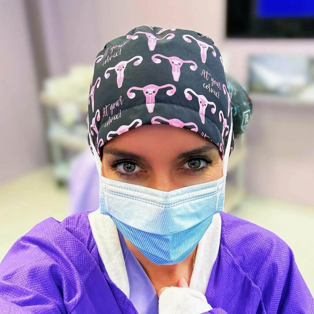 Ginecologa en quirofano con gorro quirurgico Robin Hat modelo Cervix con patron de uteros rosas y texto At your cervix sobre fondo oscuro, diseño exclusivo para ginecologia y obstetricia