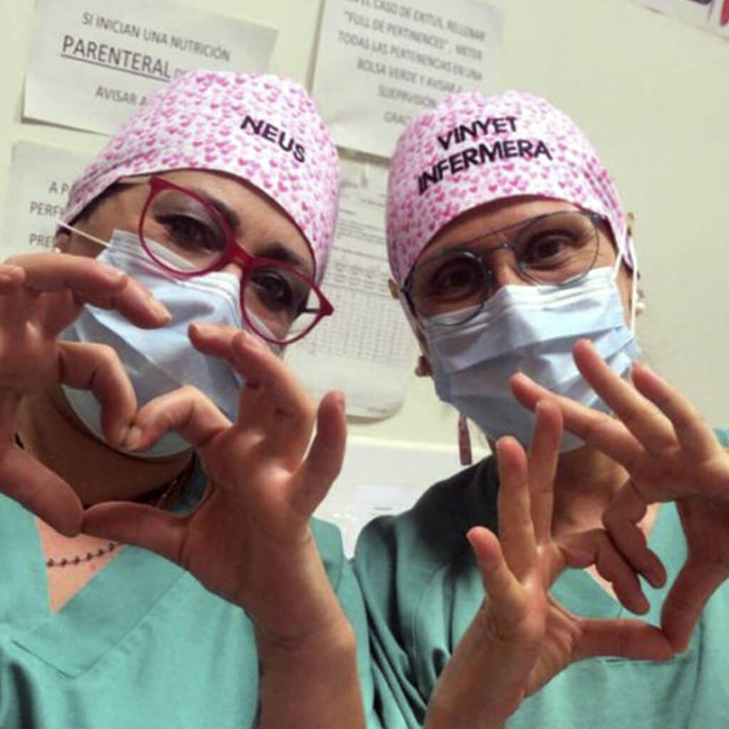 Dos enfermeras con gorros quirurgicos Robin Hat modelo Cuore personalizados con bordado de nombres NEUS y VINYET ENFERMERA, patron de corazones rosas sobre blanco, ejemplo de personalizacion Robin Custom