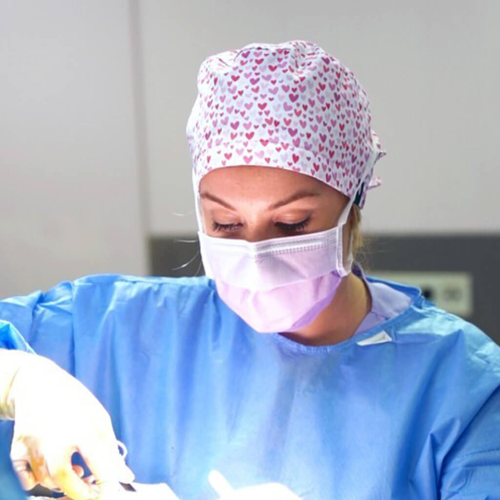 Cirujana concentrada en intervencion quirurgica con gorro Robin Hat modelo Cuore, estampado romantico de pequenos corazones rosas y morados sobre fondo blanco, ideal para profesionales con pelo largo