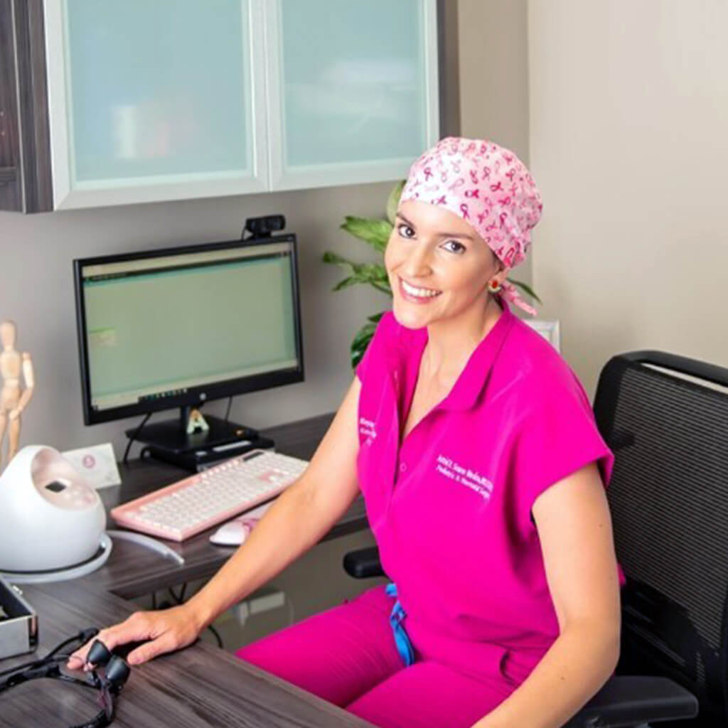Profesional sanitaria sonriente en consultorio con gorro quirurgico Robin Hat de lazos rosas awareness, simbolo de concienciacion contra el cancer de mama, uniforme medico rosa completo