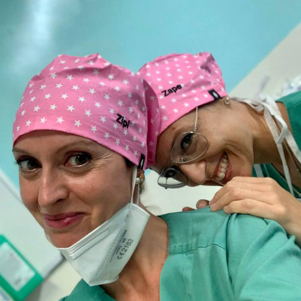 Dos profesionales sanitarias sonriendo en quirófano usando gorros quirúrgicos ESTRELLAS ROSAS de Robin Hat con estampado de estrellas blancas sobre fondo rosa, llevando mascarillas y uniformes verdes durante jornada laboral hospitalaria